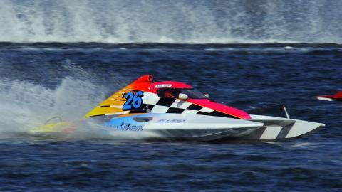 Colorful inboard hydroplane race boat numbered 26 speeding across the water, creating spray behind it during a boat race.